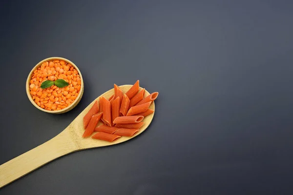 Tahta kaşıkta kırmızı makarna ve gri arka planda yuvarlak kasede kırmızı mercimek. Kırmızı Mercimek Penne Yapıştırıcısı.