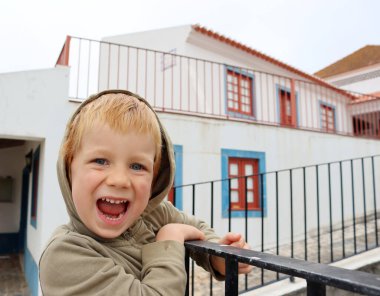 Kapüşonlu sarışın ve ağzı açık bir çocuk. Dört yaşında bir çocuk çitin yanında duruyor ve yüzünü buruşturuyor.. 