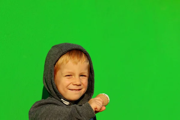 The blond hooded boy narrowed his eyes and bit his lip. A four-year-old boy makes grimaces against the background of a green wall.