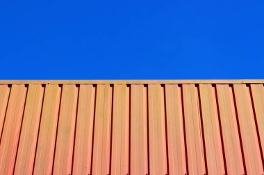 Açık mavi gökyüzüne karşı parlak turuncu kıvrımlı metal yüzeyi olan minimalist bileşim, koyu renk kontrastını ve temiz geometrik çizgileri vurguluyor..