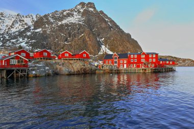 Red rorbuer-once seasonal fishing huts-now for tourist use-closed out of season-main harbor of A i Lofoten village-Tinddalstinden-Stottlikollen mounts background. Sorvagen-Moskenesoya-Nordland-Norway. clipart
