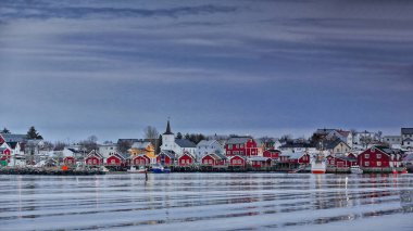 Reinevagen Körfezi 'nden Reine Köyü' ne ve liman tesislerine. Kırmızı balıkçı kulübeleri ve ufukta beyaz Reine Kirk kilisesi var. Moskenesoya-Lofoten Adaları-Nordland Fylke-Norveç.