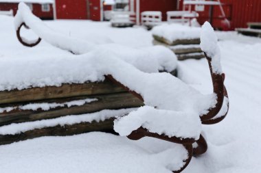 Eski paslı, beş dişli bir çapa ile karla kaplı zeminde beyaz bir kır evi-turist terörünün avlusundaki kütüklerin yanında. Hamnoy-Reine-Moskenesoya-Lofoten-Nordland-Norveç