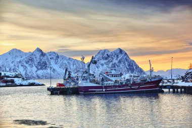 Lofoten Rorbuer süitlerinin arka plandaki liman-Litlmolla adasına demirlemiş, Svolvaer balıkçı iskelesi büyüklüğünde bordo balıkçı teknesinin üzerinde gün doğumunun olduğu NE.alandaki SE.ward manzarası. Lofoten-Nordland-Norveç.