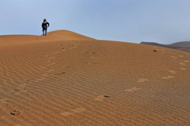 Hareket halindeki bir kum tepesindeki turist rüzgarın batıya doğru estiği tepecikleri Sumu Jaran ve Sumu Barun Jaran 'ın doğu kıyısını şekillendiren yamaçlara bakıyor. Badain Jaran Çölünün İçindeki Moğolistan-Çin.