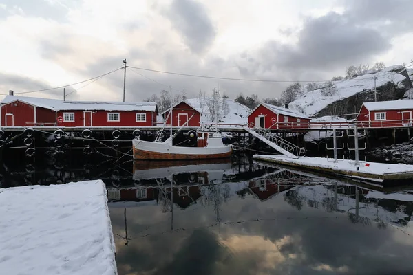 Nusfjord fishing village-snow covered harbor with floating pontoons and fishing boat moored-reflection on water-red wooden fishing huts or rorbuer for tourist use. Flakstadoya-Lofoten-Nordland-Norway.