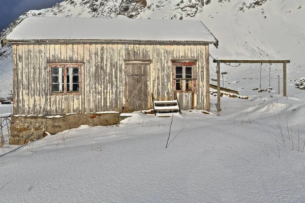 Snow covered fully ruined-abandoned-paint faded wooden hut with wooden frame of swing set-foot of snow capped mount Hustinden beside E10 European road. Vareid-Flakstadoya-Lofoten-Nordland fylke-Norway