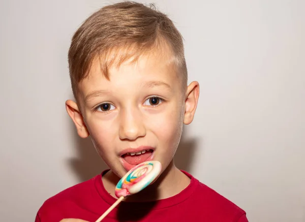 Um menino come um doce doce multicolorido em um pau, um menino com um chupa-chupa de açúcar, que ele segura em suas mãos — Fotografia de Stock