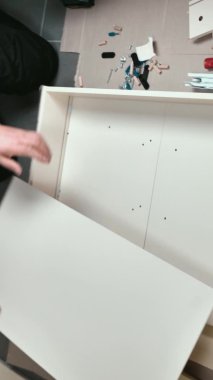 Genç bir adam evinin oturma odasında mobilya topluyor. Bir adam dolap için çekmece topluyor.