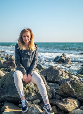 Denizin arka planında kayaların üzerinde duran bir kızın portresi.