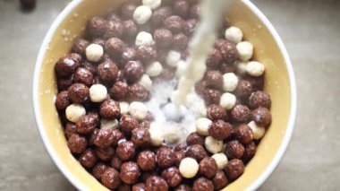 Çikolata taneli toplarla dolu bir kaseye taze süt dökülüyor. Yavaş çekimde gevrek taneciklerin gözenekli kahverengi yüzeyine sıçramış.