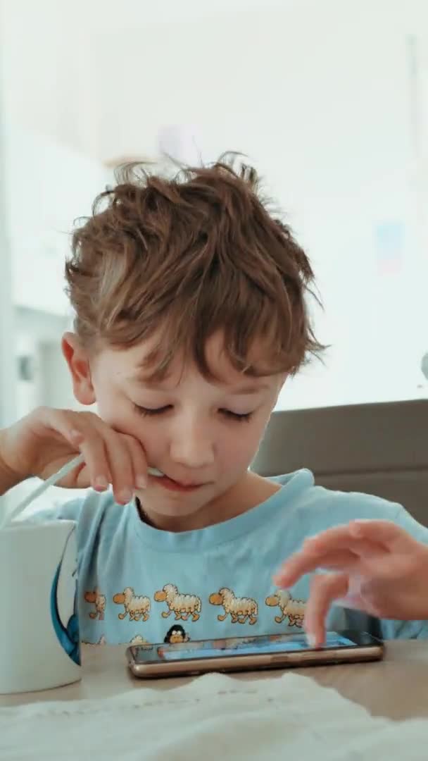 Vidéo verticale. garçon en pyjama manger le petit déjeuner à la table et regarder le téléphone — Video