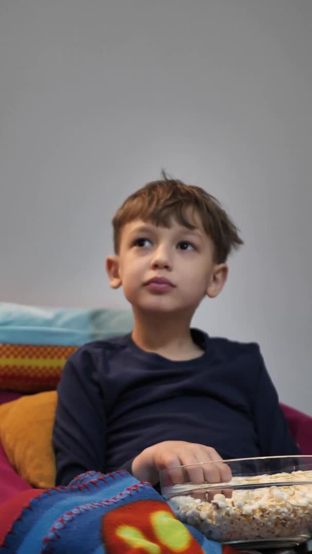 Vídeo vertical. El niño se acuesta en el sofá y come palomitas de maíz. El chico está viendo una película. Placa de palomitas de maíz en primer plano. Snack para niños — Vídeos de Stock