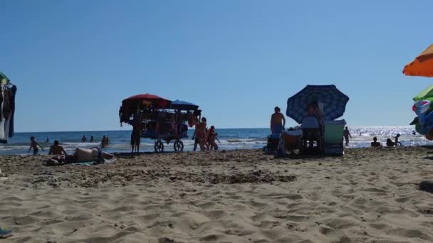 Italia, Roma 13. 06. 2021. seorang pria mencari nafkah dengan menjual pakaian di pantai, mengangkut gerobak berat dengan pakaian di bawah terik matahari — Stok Video