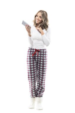 Playful feminine adorable young beauty in pajamas using cell phone. Full body length isolated on white background. clipart