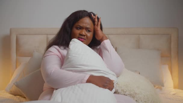 Üzgün Afrikalı kadın yatakta oturmuş yastığa sarılıyor. — Stok video