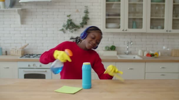 Menina adolescente africana alegre em fones de ouvido limpeza na cozinha — Vídeo de Stock