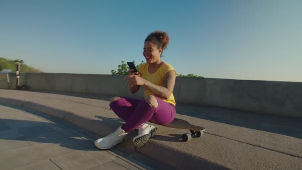 Vrolijke zwarte vrouw zit op skateboard, online messaging op telefoon — Stockvideo