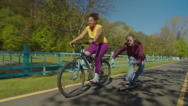 Lovely black woman cyclist speeding up female skater during skateboarding outdoors — Stock Video