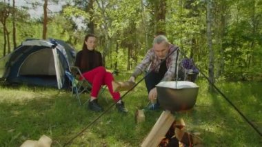 Mature camper man putting firewood in campfire during cooking