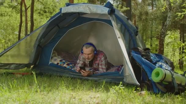 Mature hiker man in headphones enjoying music in tent on camping holiday — стоковое видео