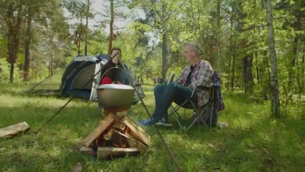 Tourist couple on camping trip drinking hot drink near campfire — стоковое видео