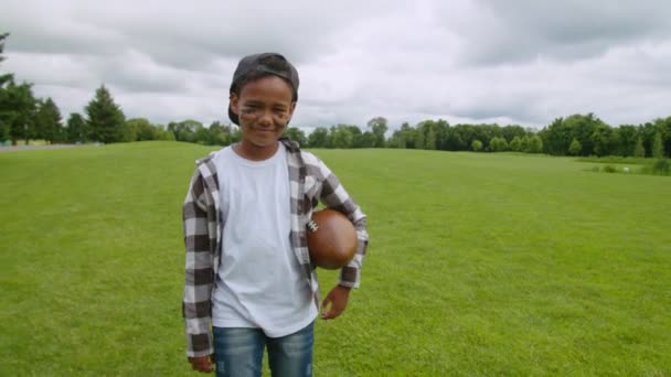 Щасливий африканський хлопчик з американським футбольним м'ячем, що ходить по полю. — стокове відео