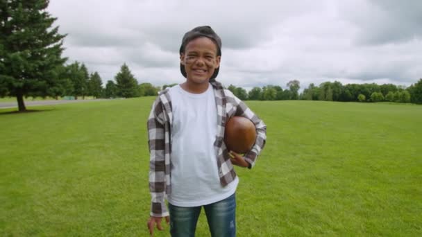 Pozitivní malý africký chlapec s americkým fotbalovým míčem pózuje venku — Stock video