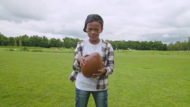 Serious little black boy holding american football ball on field — Vídeos de Stock