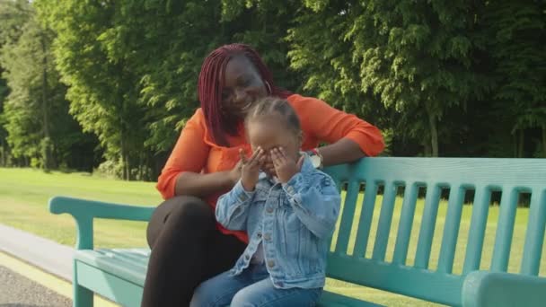 Preciosa madre africana y niña pequeña relajándose en el banco en el parque — Vídeos de Stock
