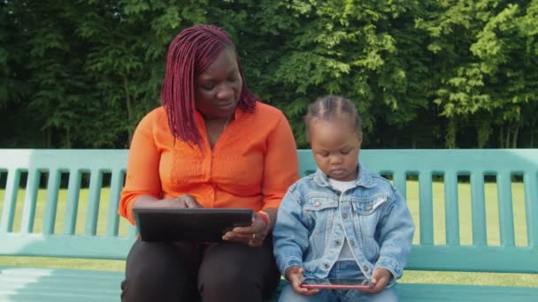 Pretty czarny matka i dziecko dziewczyna networking z cyfrowe urządzenia na ławce park — Wideo stockowe