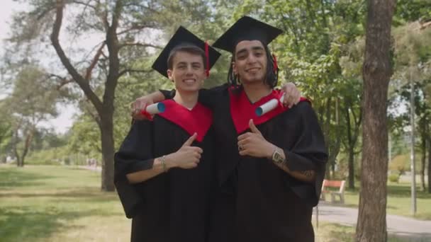 Graduados varones multiétnicos sonrientes con diplomas que muestran pulgares al aire libre — Vídeo de stock