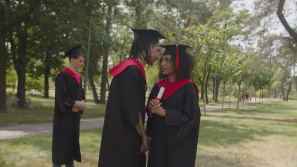 Bonito árabe macho graduado sussurrando para linda estudante negra ao ar livre — Vídeo de Stock