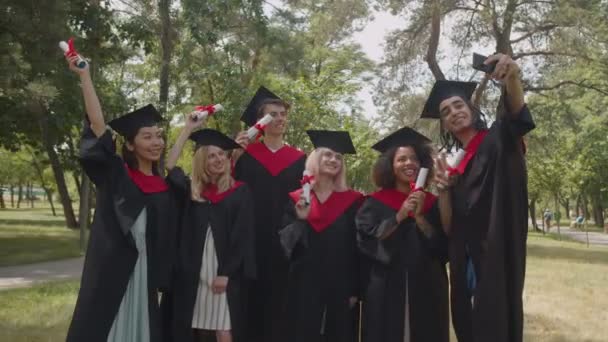 Orgullosos y diversos graduados multirraciales en vestidos académicos con diplomas tomando selfie shot — Vídeos de Stock