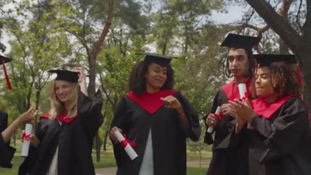 Portrét veselých tanečních multiraciálních absolventů slavících vysokoškolský titul venku — Stock video