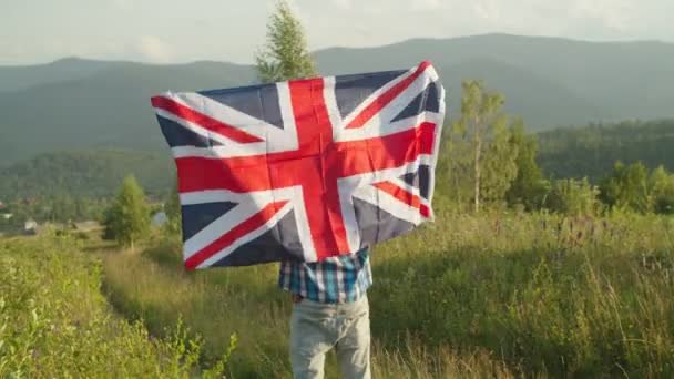 Hombre alegre de pie con gran bandera de Gran Bretaña en la cima de la montaña al atardecer — Vídeo de stock