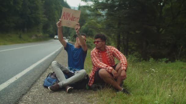 Snygga mångkulturella hipster manliga resenärer liftar med kartong på motorvägen — Stockvideo