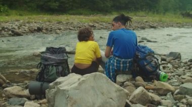 Sırt çantalı, dağ kıyısındaki kayalarda oturan romantik, çok kültürlü çift yürüyüşçülerin arka görüntüsü. Sevgi ve şefkatle kucaklaşırken, eğlence ve manzaranın tadını çıkarıyorlar.