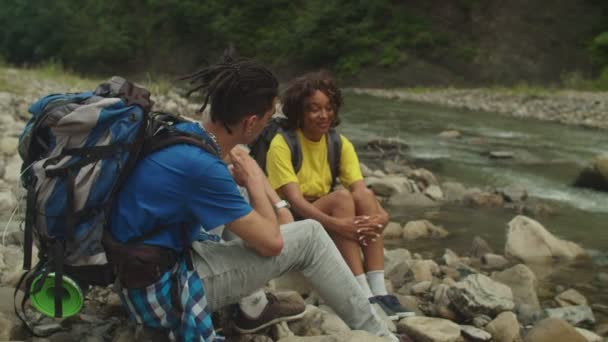 ハンサムなアラブ人男性旅行者と魅力的なアフリカ系アメリカ人女性観光客が山の川岸に岩の上に座って 山の中で多人種のカップルのバックパッカートレッキングながら話をし レジャーを楽しんでいます — ストック動画