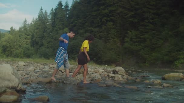 Ελκυστικό πολυπολιτισμικό ζευγάρι ταξιδιώτες wading ορεινό ποτάμι για πεζοπορία — Αρχείο Βίντεο