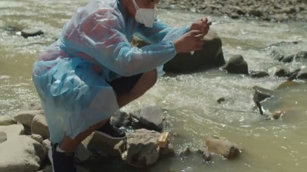 African american male scientist in protective suit sampling polluted water into test tube from river — Stock Video