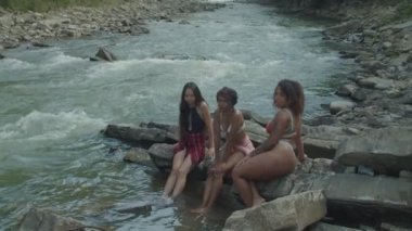 Güzel, çok kültürlü kadınlar mayo giyip kayaların üzerinde dinleniyor ve ayaklarıyla dağ nehrine sarkıyor.