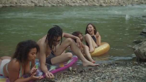 Radosni, różnorodni, wieloetniczni przyjaciele w strojach kąpielowych relaksujący się na nadmuchiwanych pierścieniach nad brzegiem rzeki — Wideo stockowe