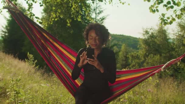 Portrait of pretty happy black woman hiker in hammock making phone call in mountains at daybreak — Stock Video