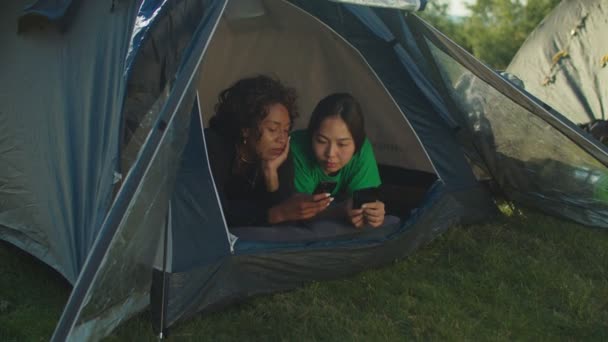 Милые многорасовые женщины-туристы, лежащие в палатке кемпинга, проверяющие фотографии по телефону во время похода в горы — стоковое видео