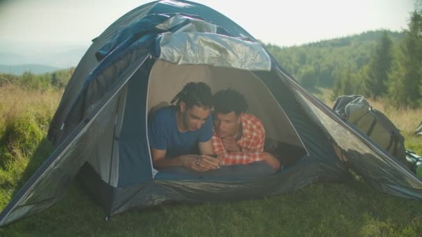Olumlu yakışıklı çok kültürlü erkek gezginler dağ yürüyüşünde kamp alanında akıllı telefonu paylaşıyorlar. — Stok video