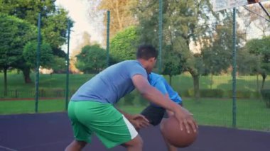 Active fit African American male streetball player driving to the hoop, passing over opposing defender and scoring two point field goals with layup shot on outdoor basketball court .