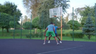 Active fit African American male streetball player performing step back shot, scoring field goals while black athletes playing half court basketball game in morning outdoors.