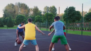 Active sporty bearded African American basketball player dribbling ball, moving past opposite defender and scoring points while diverse multiracial athletes playing streetball match on outdoor court.