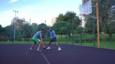 Side view of active sporty black man streetball player dribbling ball, moving past opposing defender and scoring two point field goals with layup shot while playing basketball game on outdoor court.